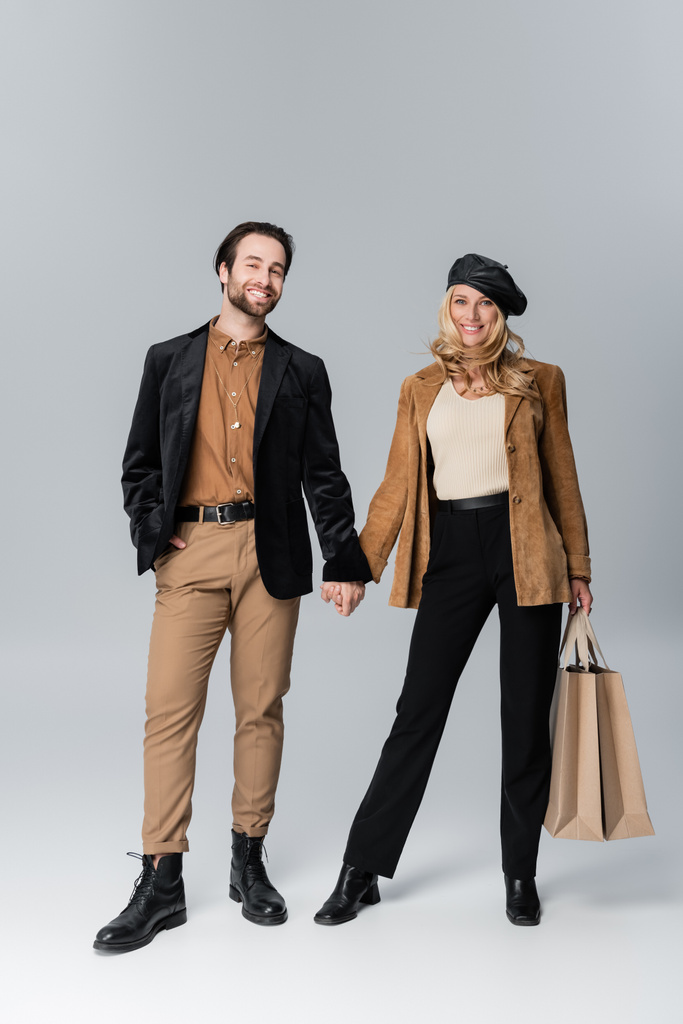 cheerful man holding hands with blonde woman in stylish beret standing with shopping bags on grey - Valokuva, kuva