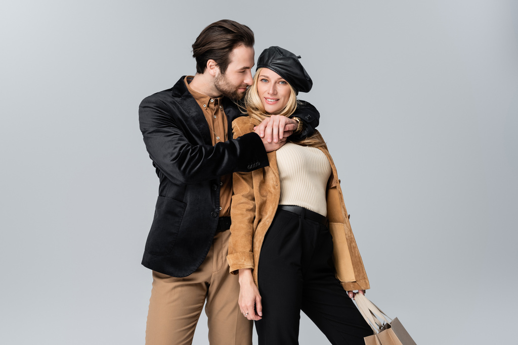 bearded man hugging happy blonde woman in stylish beret holding shopping bags isolated on grey - Foto, Imagem