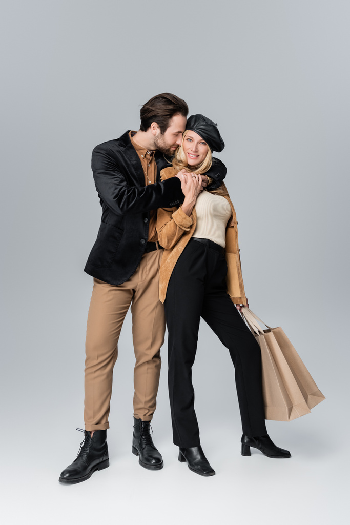 bearded man hugging happy woman in stylish beret holding shopping bags on grey - Fotografie, Obrázek