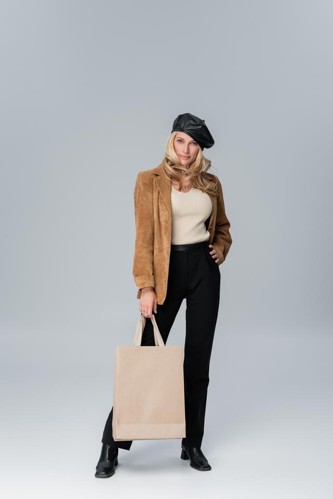 full length of blonde woman in stylish leather beret and beige blazer holding shopping bag on grey - Foto, Imagen