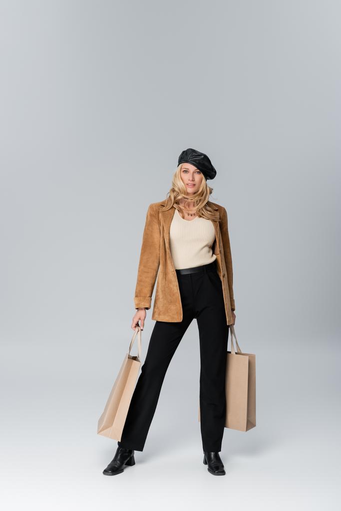 full length of happy woman in stylish leather beret and beige blazer holding shopping bags on grey - Photo, image