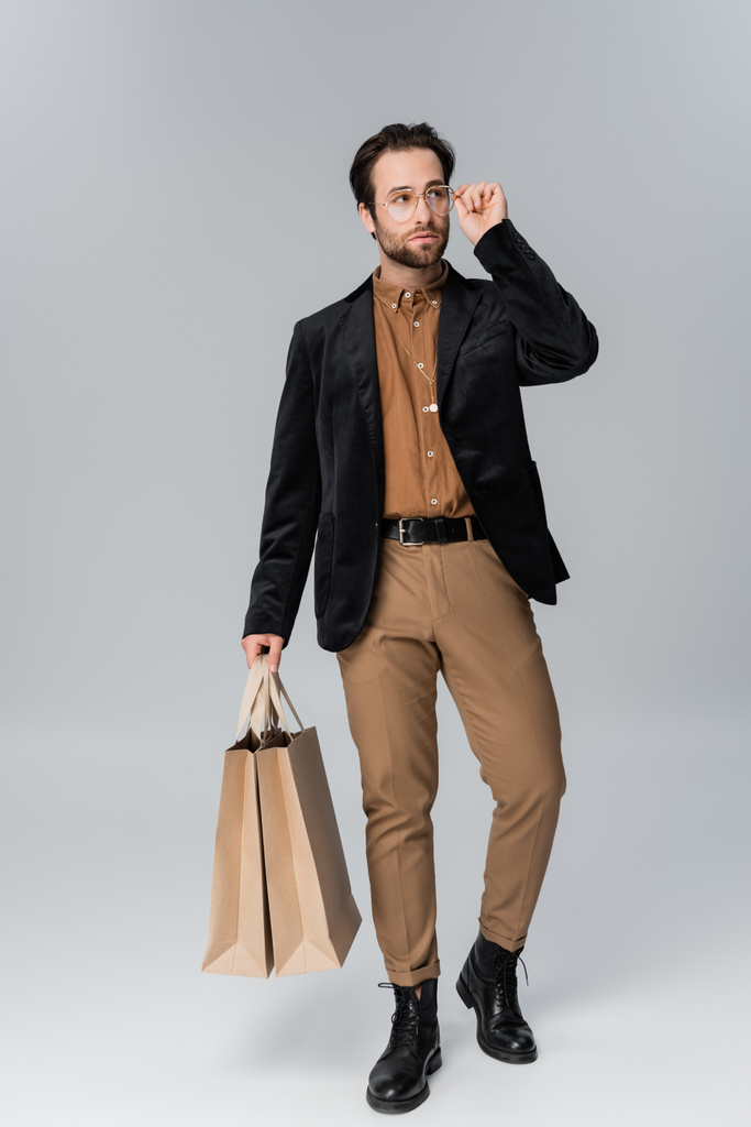 full length of bearded man in stylish autumnal outfit adjusting sunglasses and posing with shopping bags on grey - Valokuva, kuva