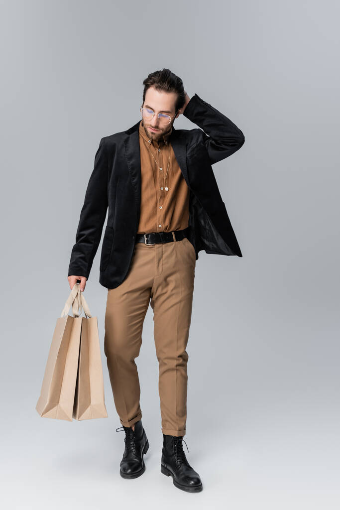 full length of bearded model in sunglasses and stylish autumnal outfit posing with shopping bags on grey - Fotoğraf, Görsel