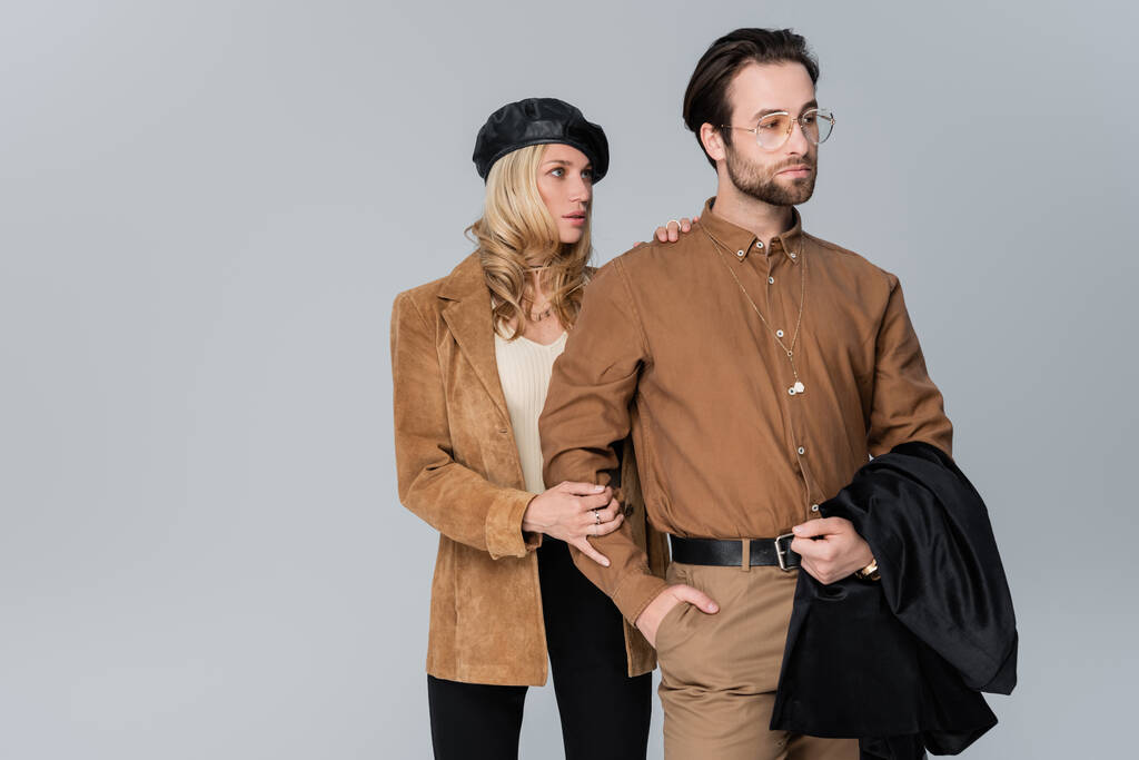 blonde woman in stylish beret hugging bearded man in sunglasses and posing with hand in pocket isolated on grey - Photo, image