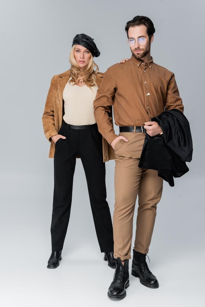 full length of blonde woman in stylish beret hugging bearded man in sunglasses and posing on grey - 写真・画像