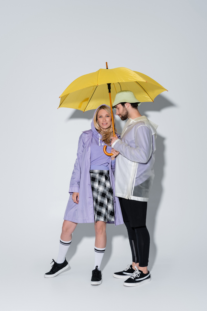 full length of woman in tartan skirt and longs socks standing with man in panama hat under yellow umbrella on grey - Foto, Imagem