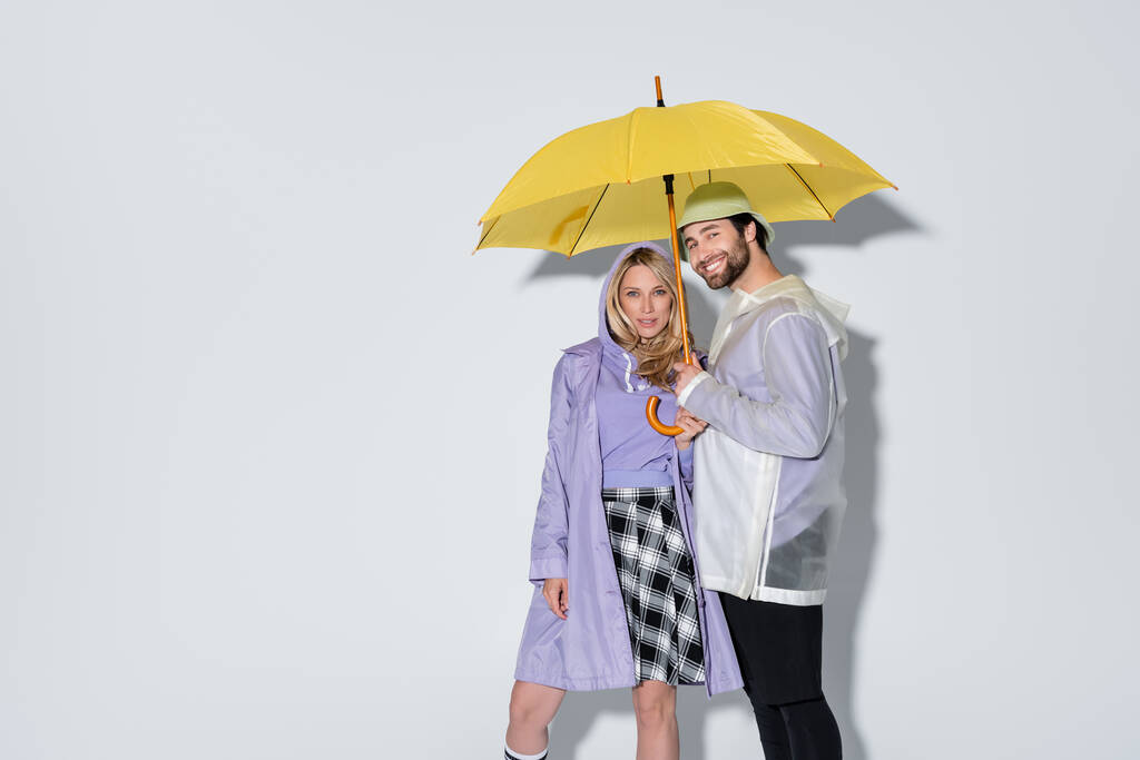 woman in tartan skirt standing with happy man in panama hat under yellow umbrella on grey - 写真・画像