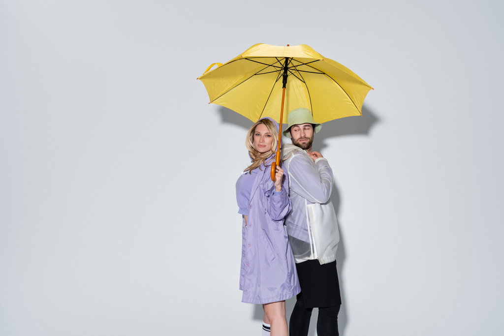 blonde woman in tartan skirt standing with bearded man in panama hat under yellow umbrella on grey - Photo, image