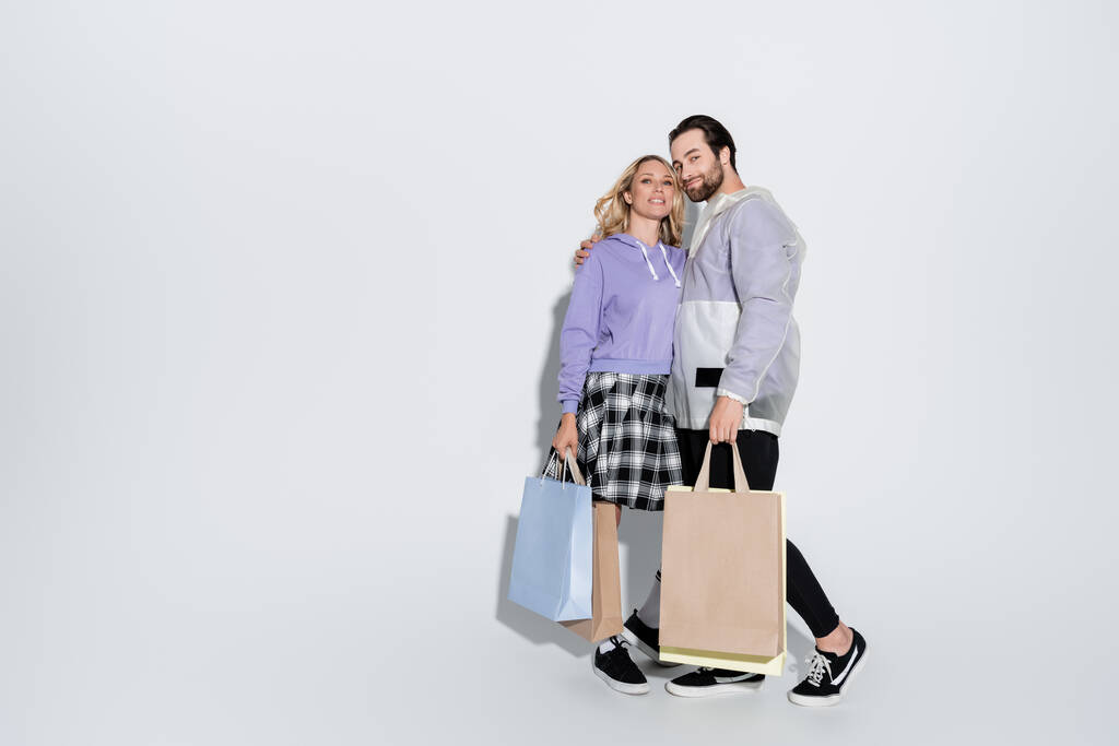full length of happy man and blonde woman in tartan skirt holding shopping bags on grey - Photo, image
