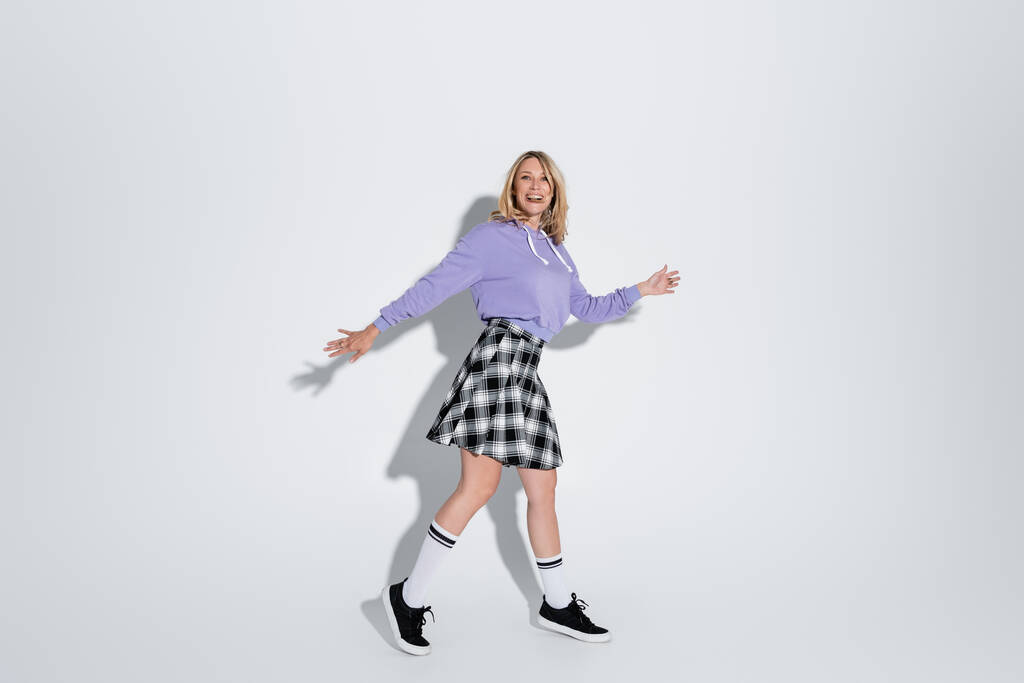 full length of excited blonde woman in trendy outfit posing with outstretched hands on grey - Foto, afbeelding
