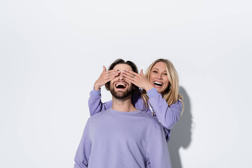 happy blonde woman covering eyes of bearded man in purple sweatshirt on grey - 写真・画像