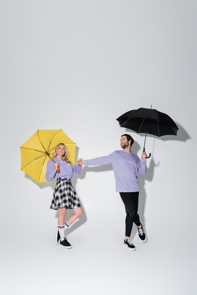 full length of joyful couple in purple sweatshirts holding hands while standing under umbrellas on grey - Photo, image