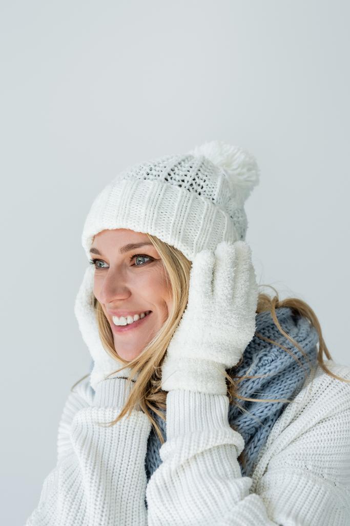 πορτρέτο της ξανθιάς γυναίκας σε χειμερινό καπέλο και λευκά γάντια αγγίζοντας το πρόσωπο και χαμογελώντας απομονωμένο σε γκρι - Φωτογραφία, εικόνα