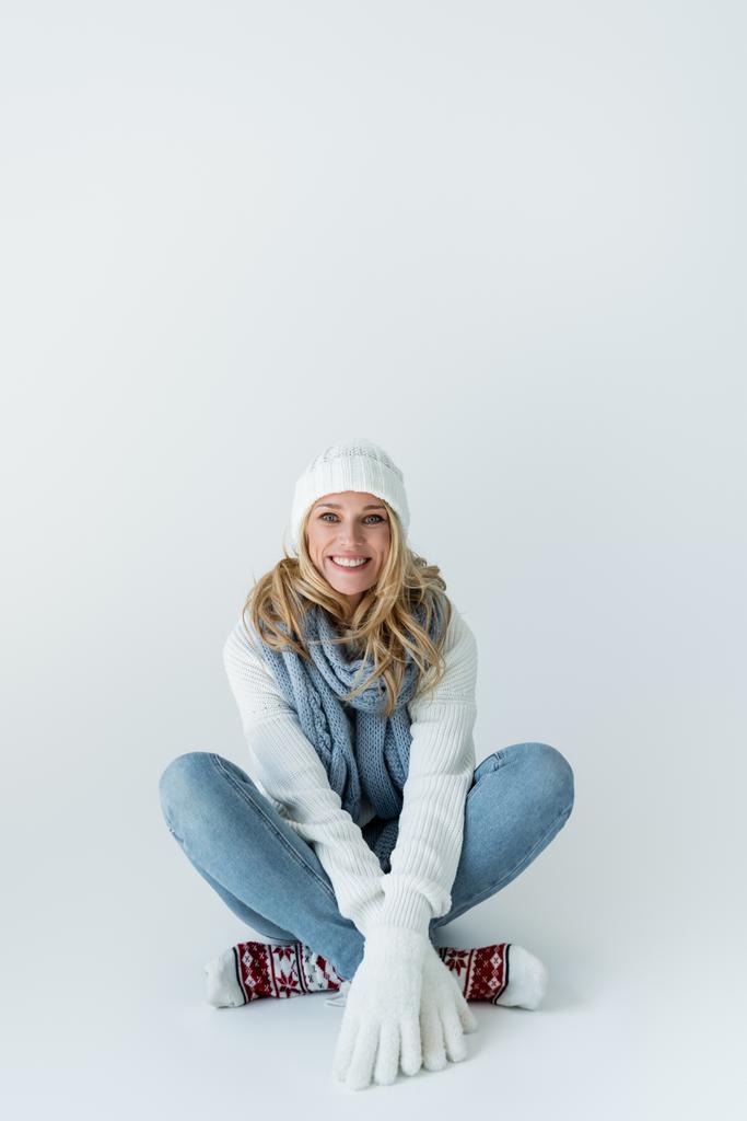 ευτυχισμένη ξανθιά γυναίκα σε χειμερινό καπέλο και πλεκτό κασκόλ κοιτάζοντας κάμερα, ενώ κάθεται σε γκρι - Φωτογραφία, εικόνα