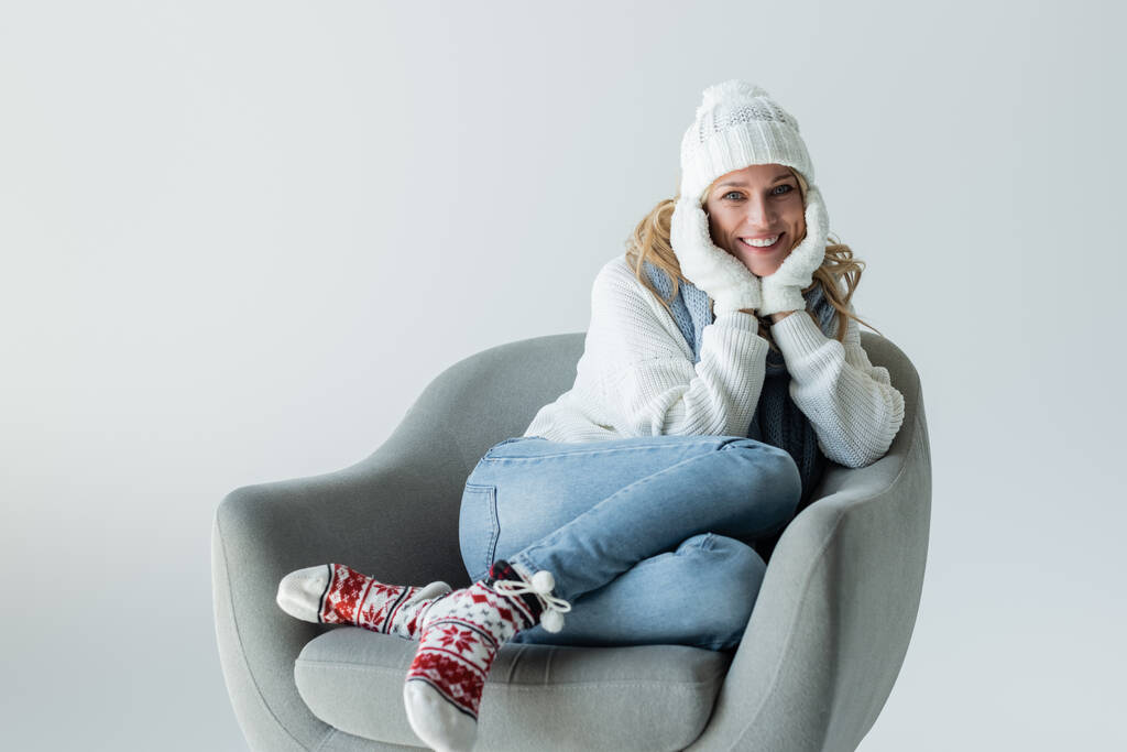 χαρούμενη ξανθιά γυναίκα με χειμωνιάτικη στολή κάθεται σε άνετη πολυθρόνα απομονωμένη σε γκρι - Φωτογραφία, εικόνα