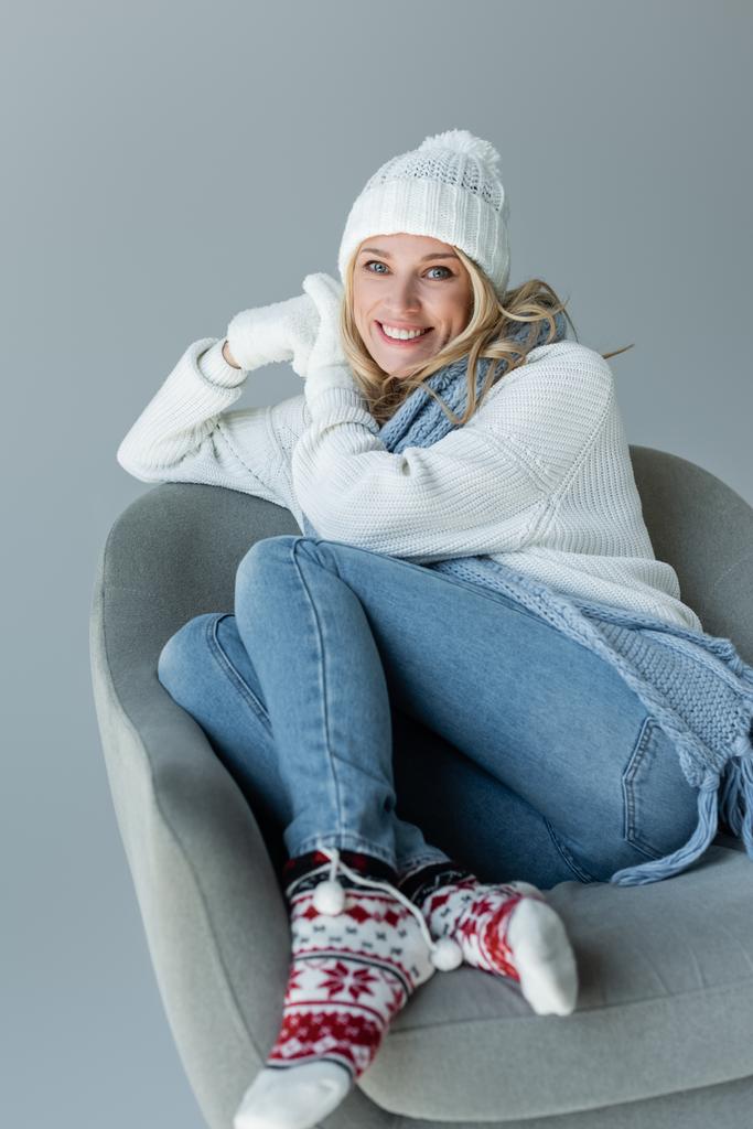cheerful blonde woman in winter outfit sitting in comfortable armchair isolated on grey - Photo, image