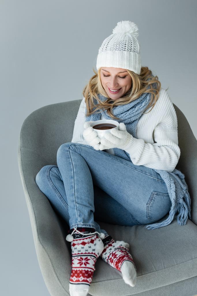cheerful blonde woman in winter outfit sitting in comfortable armchair and holding cup of coffee isolated on grey - Fotó, kép