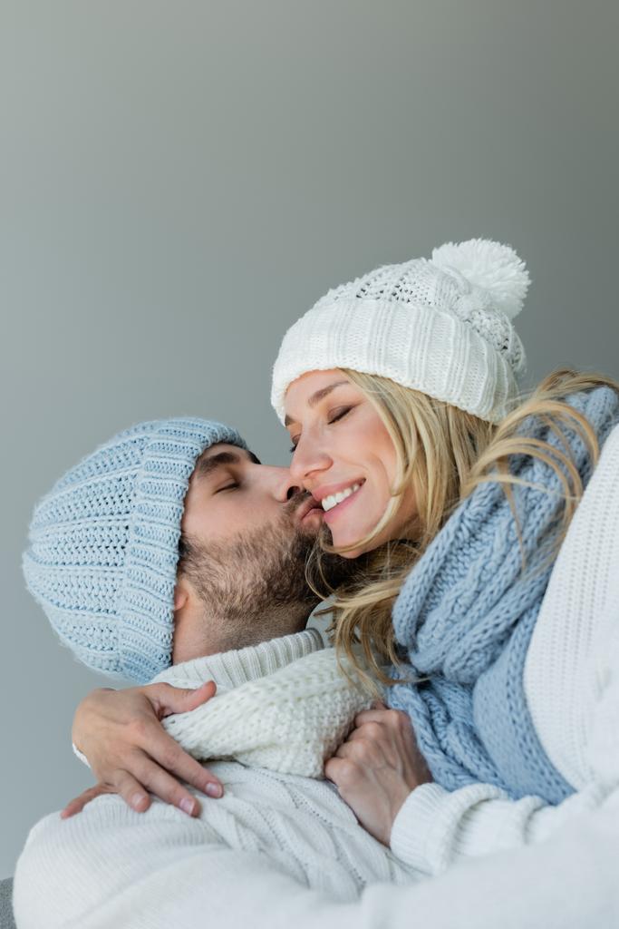 bearded man kissing cheek of happy blonde woman in knitted hat isolated on grey - Foto, Imagen