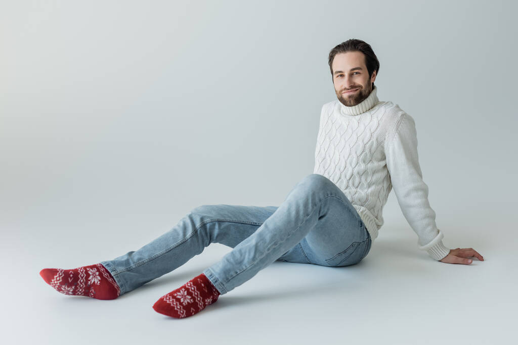 повна довжина бородатого чоловіка в джинсах і білому светрі, сидячи в червоних шкарпетках з орнаментом на сірому
  - Фото, зображення