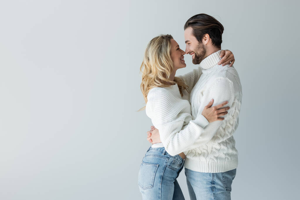side view of cheerful couple in knitted white sweaters and jeans smiling while hugging isolated on grey - Foto, afbeelding