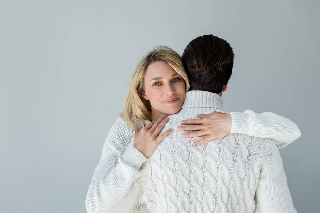 blonde woman in white sweater hugging back of boyfriend and smiling isolated on grey - Fotografie, Obrázek