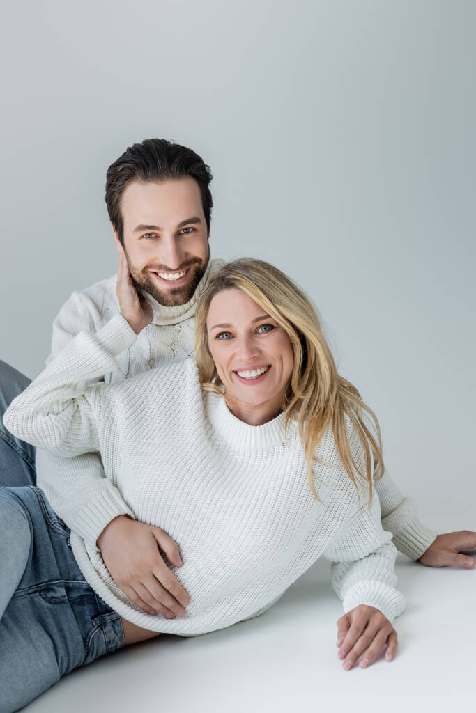 счастливые мужчина и женщина в белых вязаных свитерах смотрят в камеру на сером - Фото, изображение