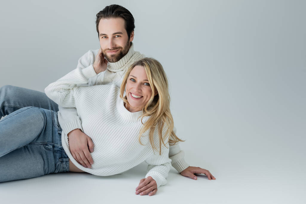cheerful man and woman in white knitted sweaters looking at camera on grey - Foto, imagen