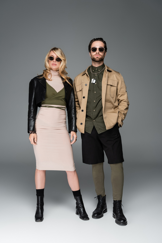 full length of stylish couple in fashionable outfits and sunglasses posing together on grey - Foto, Bild