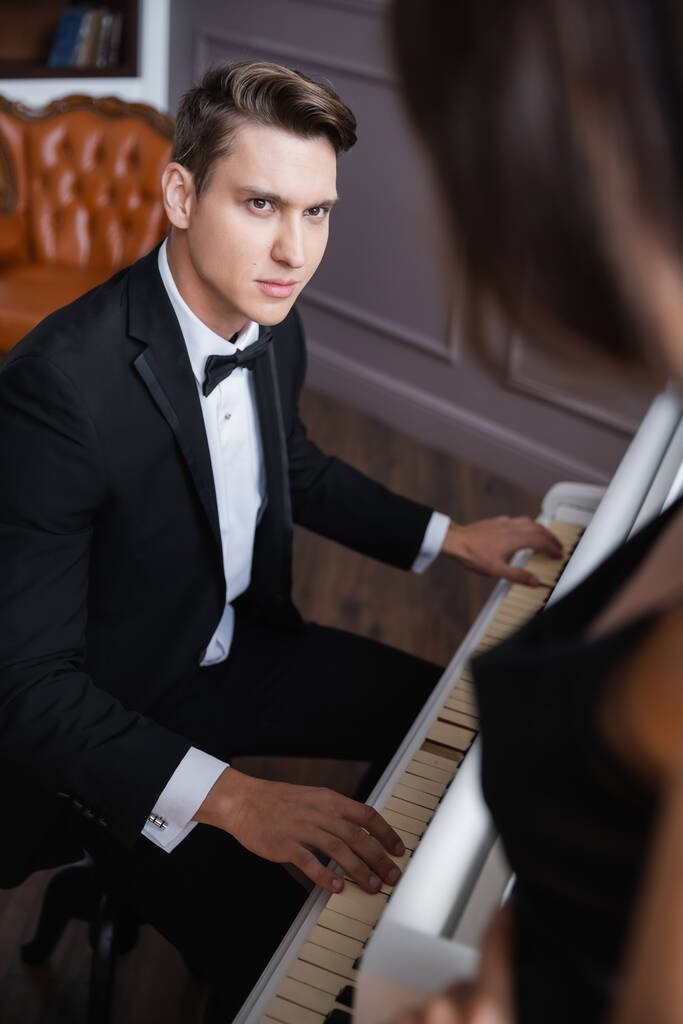 Κομψός άντρας με κοστούμι παίζει πιάνο κοντά σε θολή φιλενάδα στο σπίτι - Φωτογραφία, εικόνα