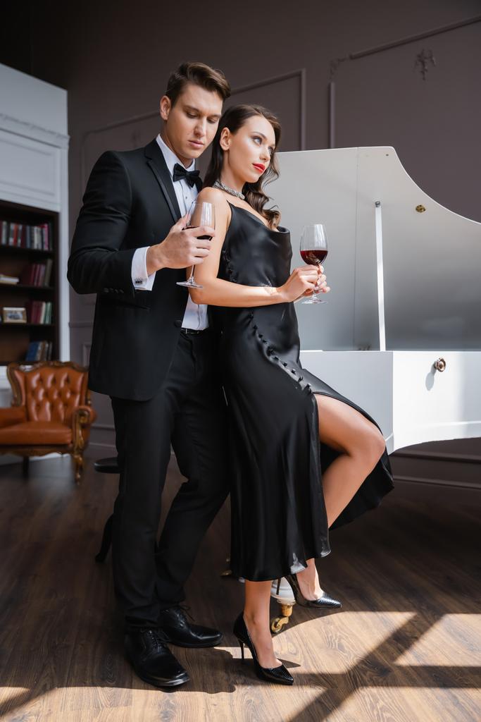 Elegant man in suit holding glass of wine near girlfriend and piano at home  - Foto, imagen