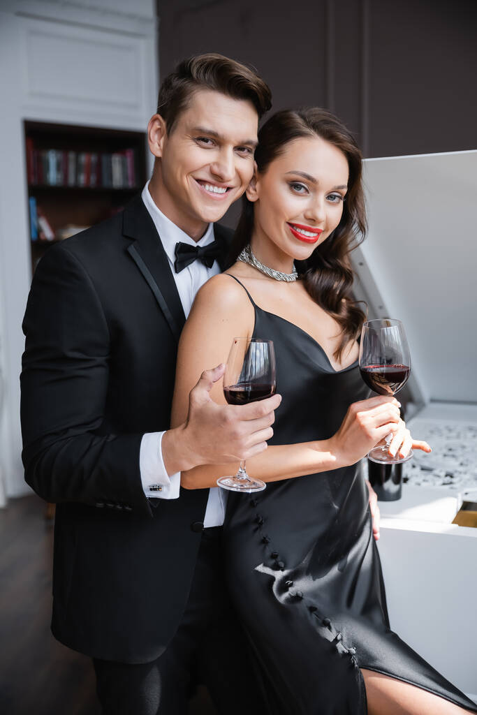Κομψό ζευγάρι κρατώντας ποτήρια κρασί και κοιτάζοντας την κάμερα στο σπίτι  - Φωτογραφία, εικόνα
