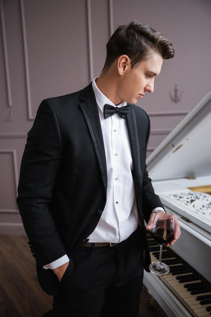 Side view of elegant man in suit holding glass of wine near piano at home  - Photo, image