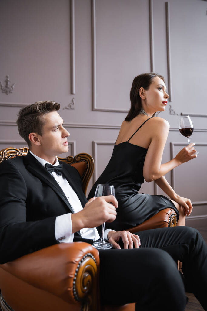 Elegant woman holding wine while sitting near boyfriend on armchair at home  - Zdjęcie, obraz