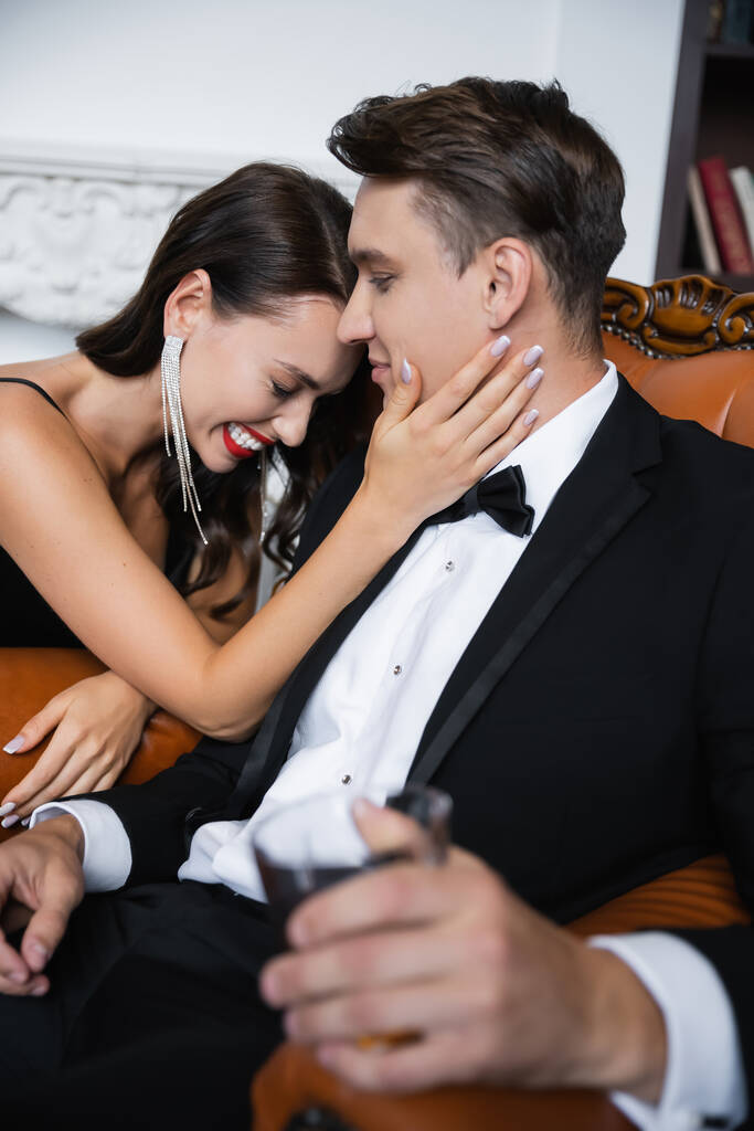 Cheerful woman with red lips touching boyfriend in suit holding blurred glass at home  - Photo, image