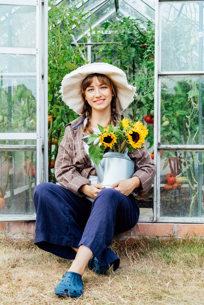 Portrait of smiling young female farmer woman holding watering can with fresh sunflowers bouquet, sitting near greenhouse. Cottagecore lifestyle. Growing organic vegetables in garden. Vertical card. - Foto, Imagem