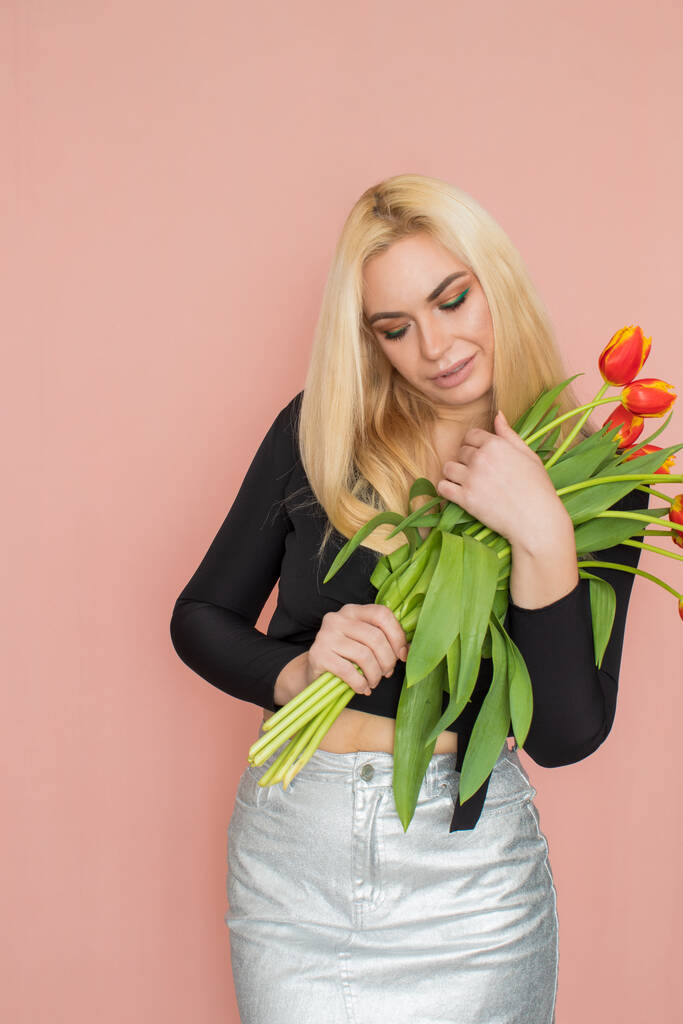 Fashion model woman in fashionable clothes on pink background. Wearing stylish clothing, black blouse, silver skirt. Posing in studio. Holding red tulips in her hands - Fotoğraf, Görsel
