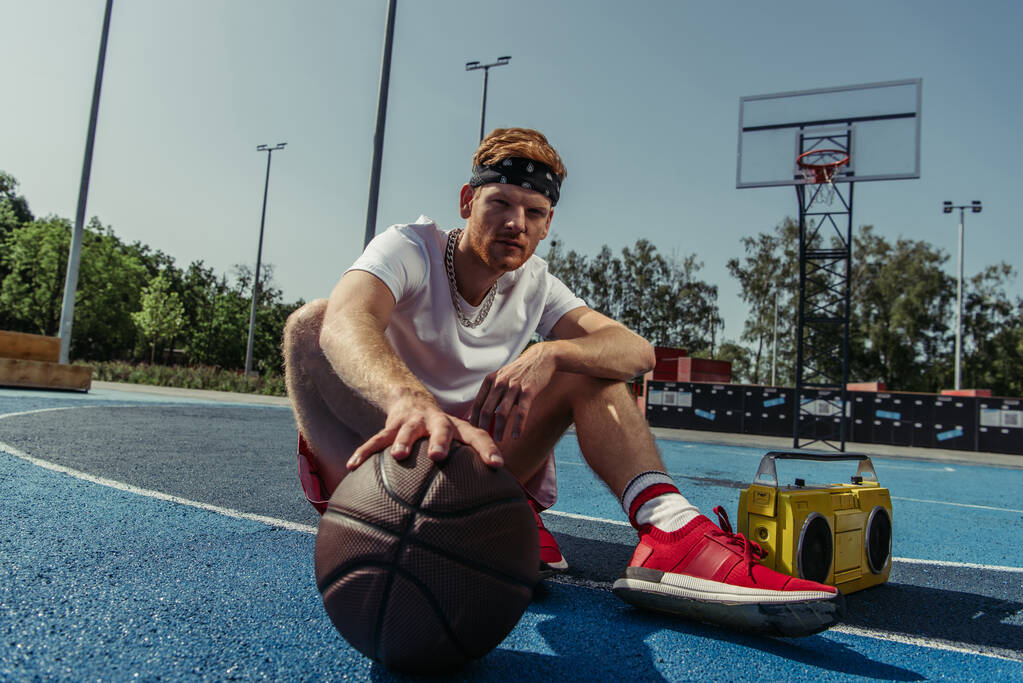 баскетболіст у спортивному одязі та бандані сидить на корті з м'ячем та магнітофоном
 - Фото, зображення