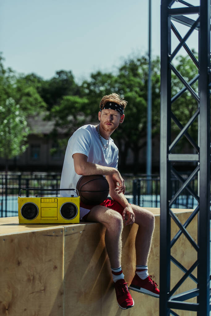 full length of man with ball sitting near boombox and looking at camera - Photo, Image