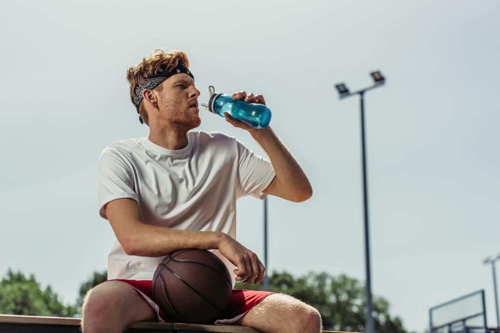 νεαρός μπασκετμπολίστας πόσιμο νερό από αθλητικό μπουκάλι, ενώ κάθεται σε εξωτερικούς χώρους - Φωτογραφία, εικόνα
