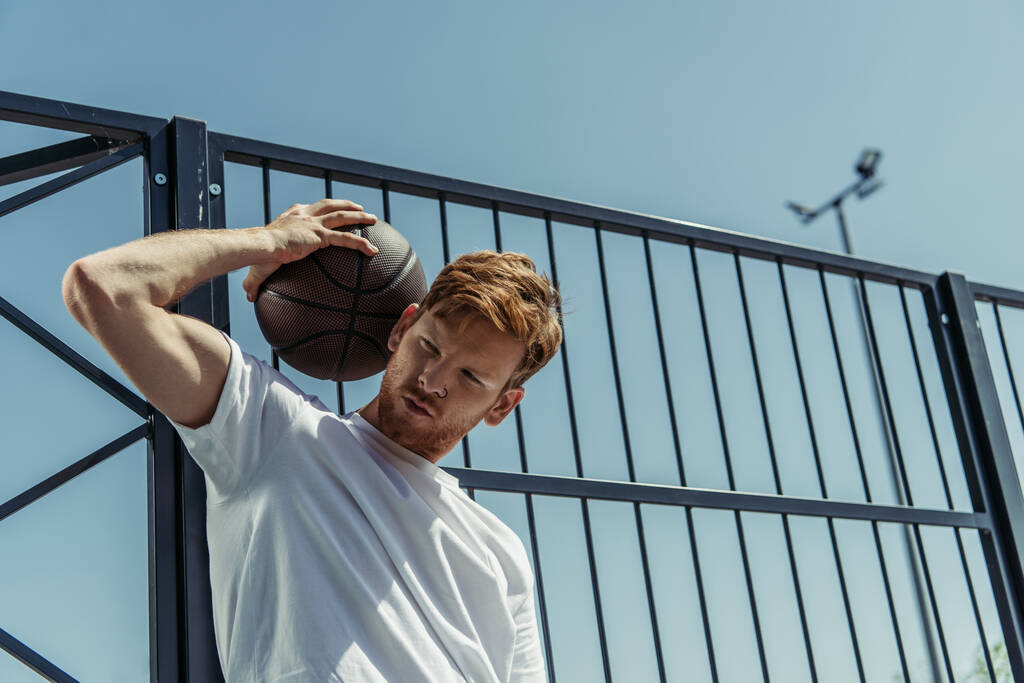 low angle view of redhead man in white t-shirt standing with ball near fence  - Zdjęcie, obraz