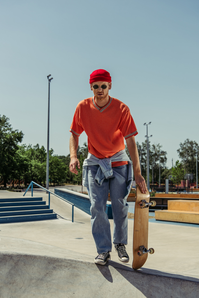 full length of man in jeans and orange t-shirt holding skateboard in park - Photo, Image