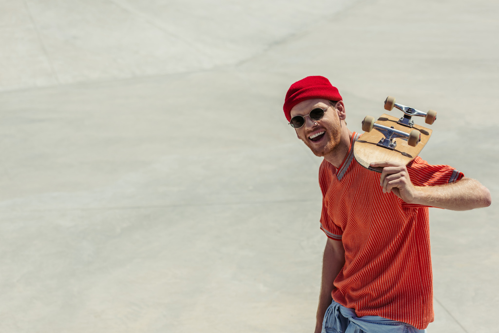 excited man in red beanie and orange t-shirt holding skateboard and looking at camera - Foto, Imagem