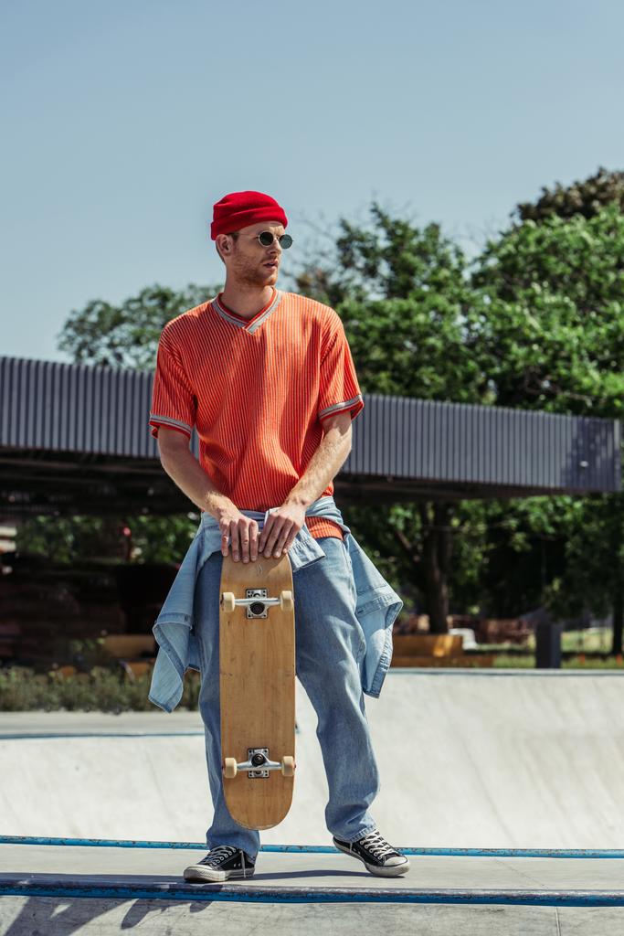 full length of fashionable man with skateboard standing in park - Fotoğraf, Görsel