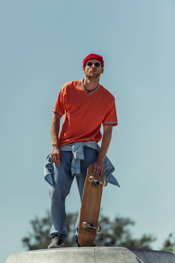 low angle view of man in sunglasses and orange t-shirt holding skateboard outdoors - Fotografie, Obrázek