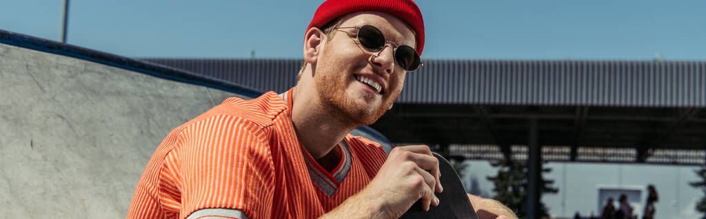 trendy skateboarder in red beanie and sunglasses smiling outdoors, banner - Valokuva, kuva