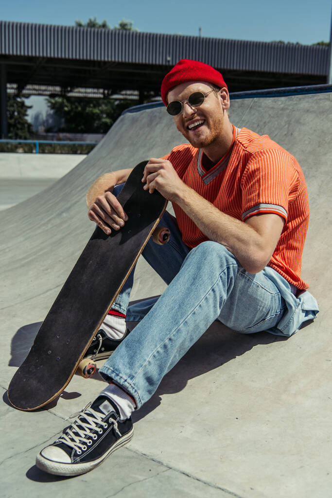 happy and fashionable man sitting with skateboard and smiling at camera - Photo, Image