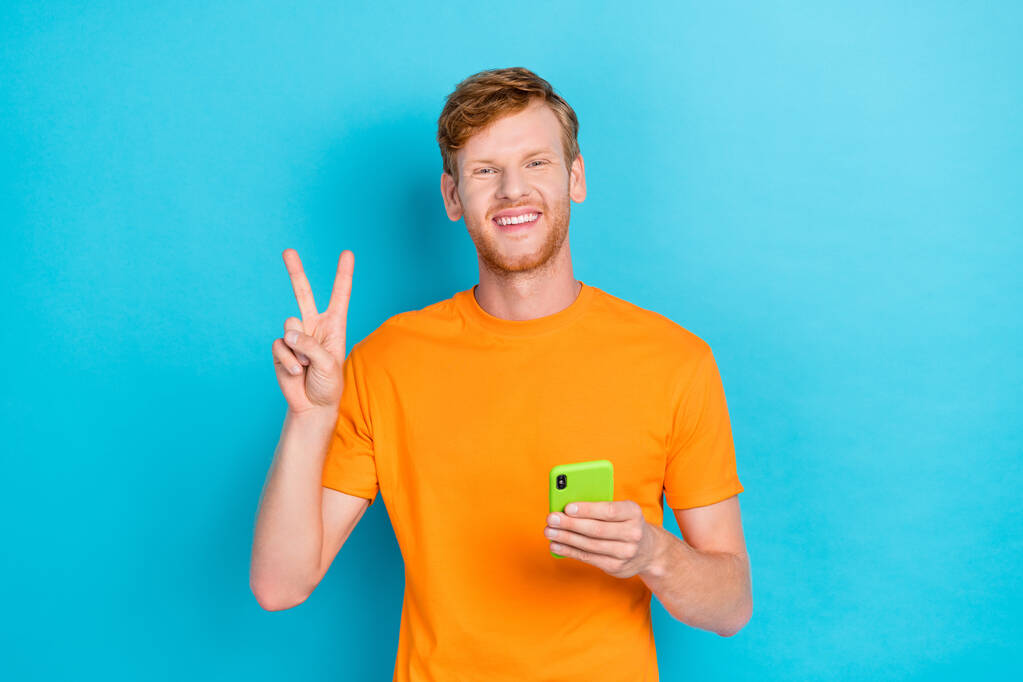 Portrait of positive cheerful handsome guy with red hairstyle wear orange t-shirt showing v-sign isolated on turquoise color background. - Photo, image