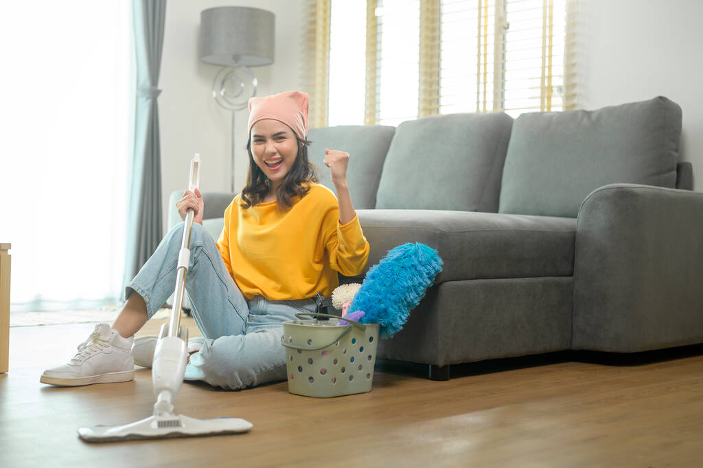 Μια νεαρή ευτυχισμένη γυναίκα σφουγγαρίζει και σκουπίζει το πάτωμα στο σαλόνι.. - Φωτογραφία, εικόνα