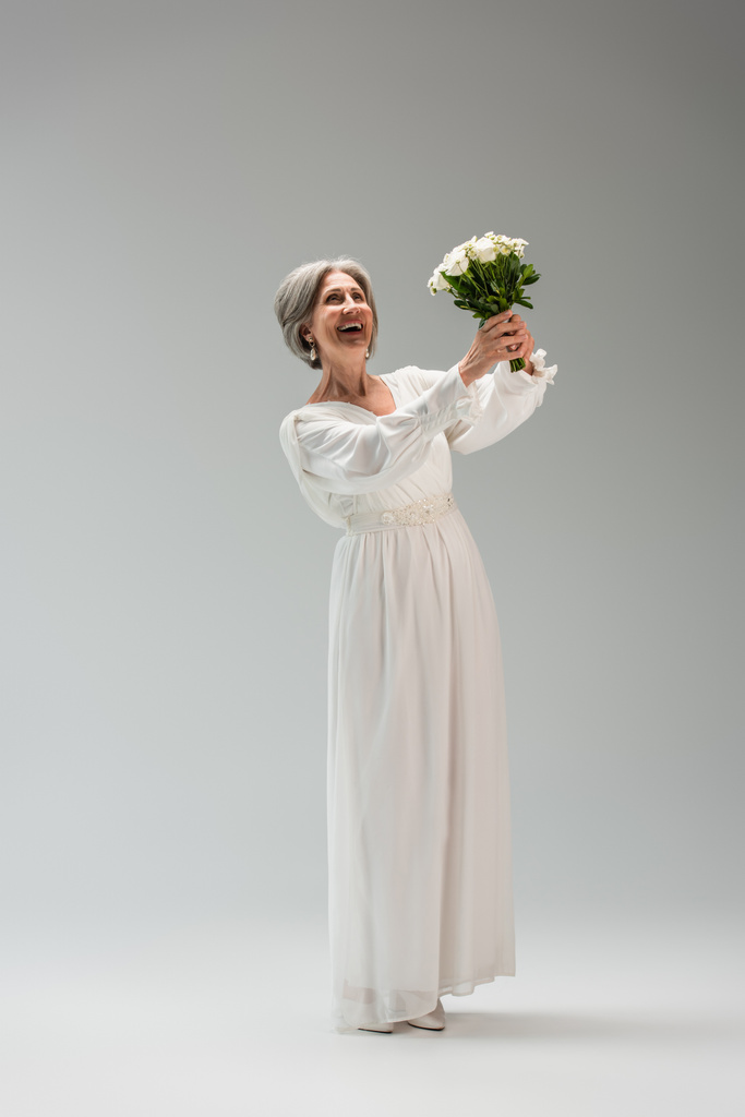 πλήρες μήκος του ενθουσιασμένοι μεσήλικες νύφη σε λευκό φόρεμα κρατώντας γαμήλιο μπουκέτο σε γκρι - Φωτογραφία, εικόνα