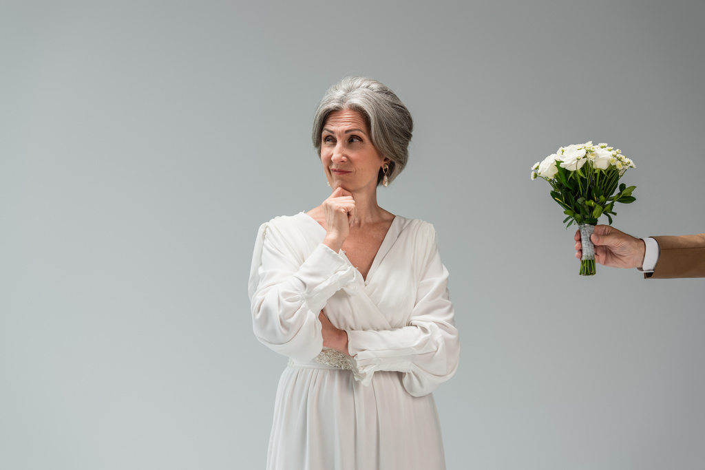 groom holding wedding bouquet near puzzled middle aged bride in white dress isolated on grey - Foto, Imagem